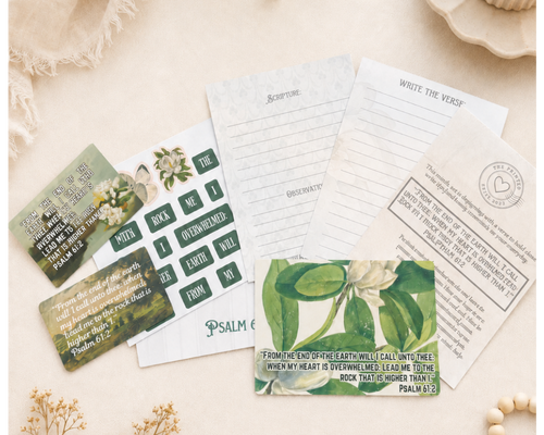 A styled flat-lay of a Bible study or prayer card set on a warm cream fabric background, surrounded by dried flowers, a lit candle, and wooden beads. The set includes printed cards with scripture verses, a writing guide, and a green botanical-themed card.